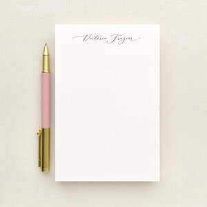 May include: A rectangular white notepad with the name Victoria Frazier printed in a dark script font at the top, positioned next to a ballpoint pen featuring a matte pink barrel and a metallic gold cap, clip, and tip.