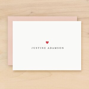 May include: A white stationery card with a red heart and the name "JUSTINE ADAMSON" printed in black. The card is inside a light pink envelope, set against a light wood surface.