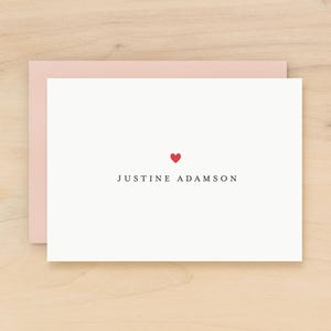May include: A white stationery card with a red heart and the name "JUSTINE ADAMSON" printed in black. The card is inside a light pink envelope, set against a light wood surface.