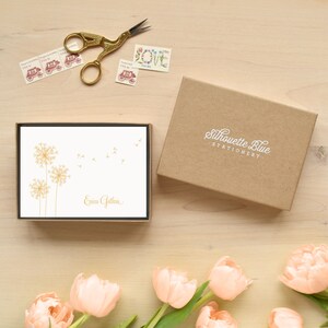 May include: A box of personalized stationery with a dandelion design in gold. The box is brown and has the text "Silhouette Blue Stationery" printed on it. The stationery is white with the name "Erica Guthrie" printed in gold.