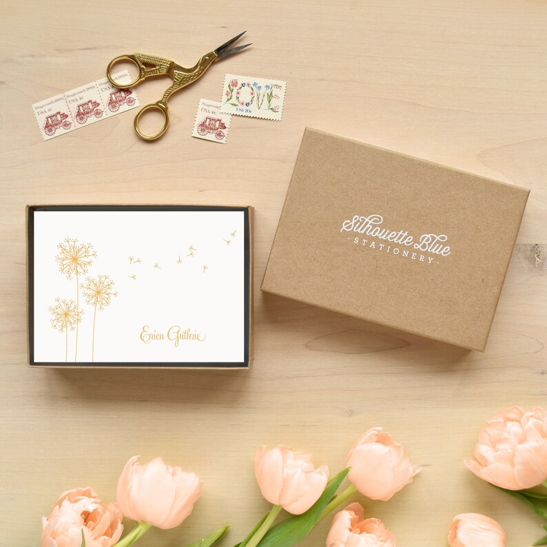 May include: A box of personalized stationery with a dandelion design in gold. The box is brown and has the text "Silhouette Blue Stationery" printed on it. The stationery is white with the name "Erica Guthrie" printed in gold.