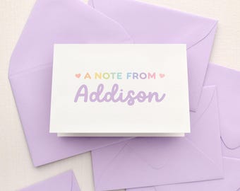 May include: A white folded stationery card with the text A NOTE FROM in a rainbow gradient font, flanked by two small pink hearts, and the name Addison printed in a large purple script font, resting on several light purple envelopes.