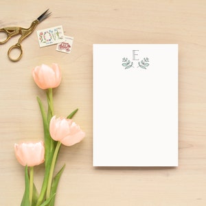 K&ouml;nnte beinhalten: Ein wei&szlig;er Notizblock mit einem schlichten Blumendesign und dem Buchstaben "E" in der Mitte. Der Notizblock liegt auf einer hellbraunen Holzoberfl&auml;che mit drei rosa Tulpen.
