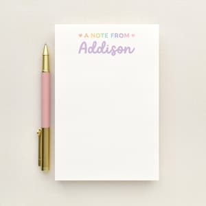May include: A white rectangular notepad with the text A NOTE FROM Addison in pastel colors, featuring small hearts on either side of the text. A pink pen with a gold clip and tip is positioned vertically along the left side of the notepad.