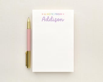 May include: A white rectangular notepad with the text A NOTE FROM Addison in pastel colors, featuring small hearts on either side of the text. A pink pen with a gold clip and tip is positioned vertically along the left side of the notepad.