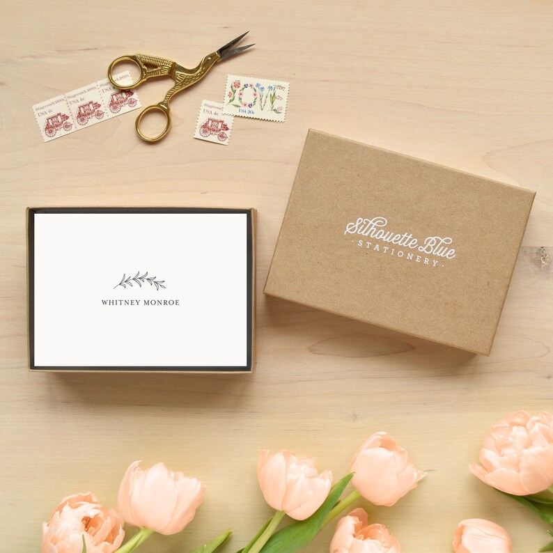 May include: A box of blank stationery cards with a simple floral design and the text "Whitney Monroe" printed on the front. The box is open and the cards are visible. The box is made of brown cardboard and has the text "Silhouette Blue Stationery" printed on the front.