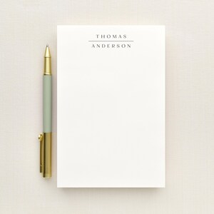 May include: A white rectangular notepad featuring the name THOMAS ANDERSON printed in black capital letters with a horizontal line above the name, accompanied by a pen with a sage green barrel and gold metallic accents on the cap and clip.
