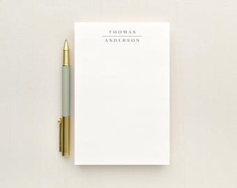 May include: A white rectangular notepad featuring the name THOMAS ANDERSON printed in black capital letters with a horizontal line above the name, accompanied by a pen with a sage green barrel and gold metallic accents on the cap and clip.