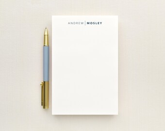 May include: A rectangular white notepad featuring the text ANDREW | MOSLEY in dark blue capital letters at the top, positioned next to a retractable pen with a light blue barrel and a metallic gold tip, clip, and end cap.