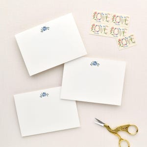 May include: Three white rectangular note cards featuring a small blue floral illustration at the top center. Accompanying the cards are five postage stamps with the word LOVE formed by floral wreaths and a pair of gold-toned metal scissors.