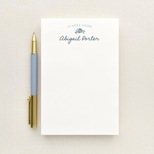 May include: A white rectangular notepad featuring the text A NOTE FROM and the name Abigail Porter in dark blue script, accented by a small blue floral illustration. A light blue pen with a gold clip and tip sits beside the notepad.
