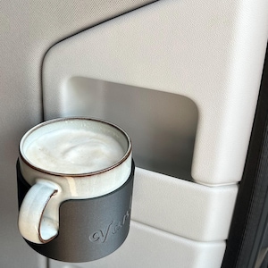 May include: A black cup holder with the word "Van" printed on it, holding a white ceramic mug with a brown rim filled with a light brown beverage.