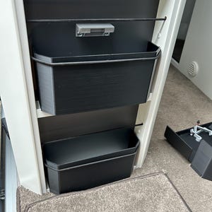 Może przedstawiać: Dwa czarne plastikowe pojemniki do przechowywania z czarną gumką, która je trzyma w miejscu. Pojemniki są zamontowane na białej ścianie z szarym metalowym zatrzaskiem.