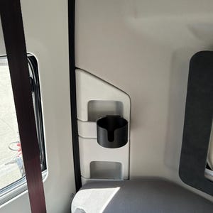 May include: A black cup holder is mounted on the side of a white interior of a vehicle. The cup holder is made of plastic and has a simple design.