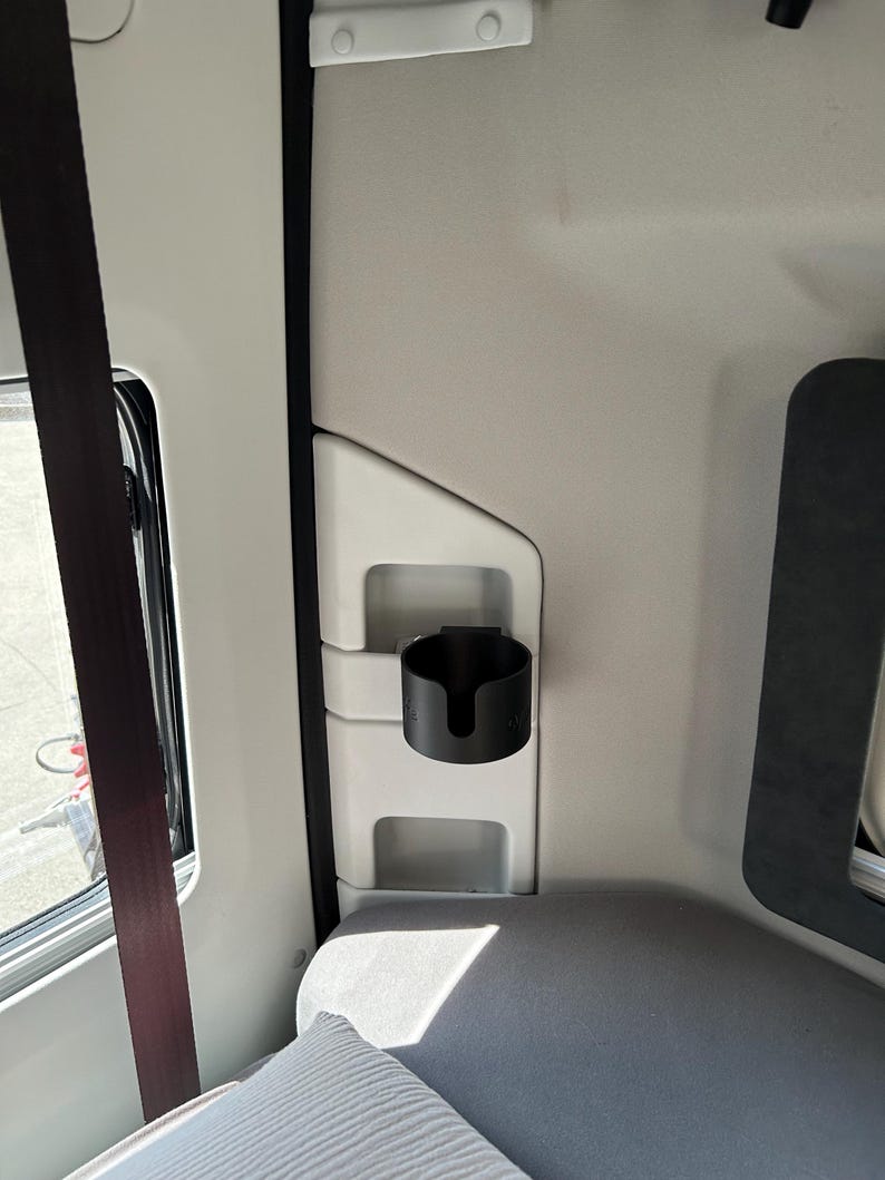 May include: A black cup holder is mounted on the side of a white interior of a vehicle. The cup holder is made of plastic and has a simple design.