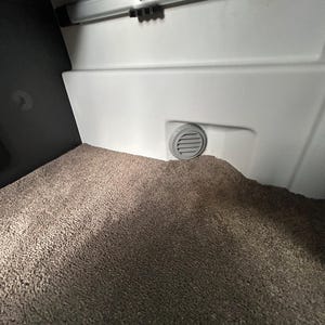 Könnte beinhalten: Innenansicht eines Fahrzeugfachs mit einer hellgrauen Kunststoffplatte, einer Lüftungsöffnung und einem strukturierten, braunen Teppichboden. Oben am Paneel ist eine Metallstange zu sehen.