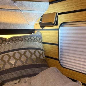 Peut inclure: Une petite étagère murale noire supporte un smartphone doré relié à un câble de charge blanc. L'étagère est fixée sur un panneau mural à motif bambou, à côté d'un store plissé, d'un oreiller à motifs et d'une couverture grise.