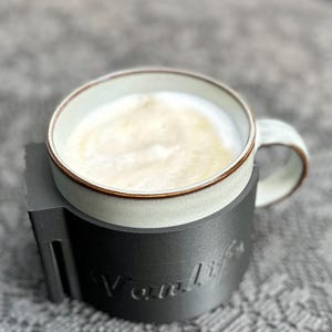May include: A white ceramic mug with a brown rim filled with coffee and a black 3D printed mug holder with the word "Vanlife" printed on it.