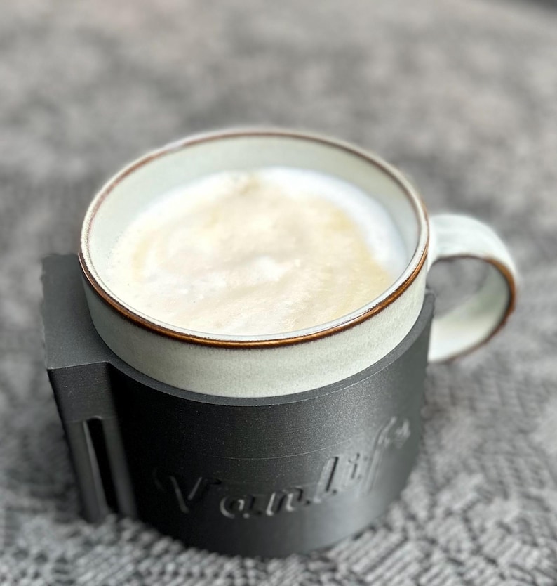 May include: A white ceramic mug with a brown rim filled with coffee and a black 3D printed mug holder with the word "Vanlife" printed on it.