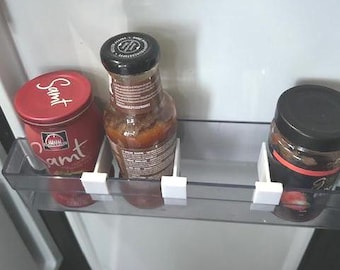 May include: A clear plastic refrigerator door shelf organizer featuring three white 3D-printed dividers. The shelf holds a red jar labeled Samt, a glass bottle with a black cap, and a dark jar with a black lid, all separated by the white clips.