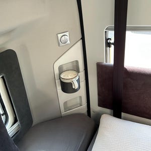 May include: A white ceramic mug with a black handle sits in a holder on the wall of a truck cab. The mug is filled with a light brown beverage. The cab has a gray and white interior with a window and a gray bed.