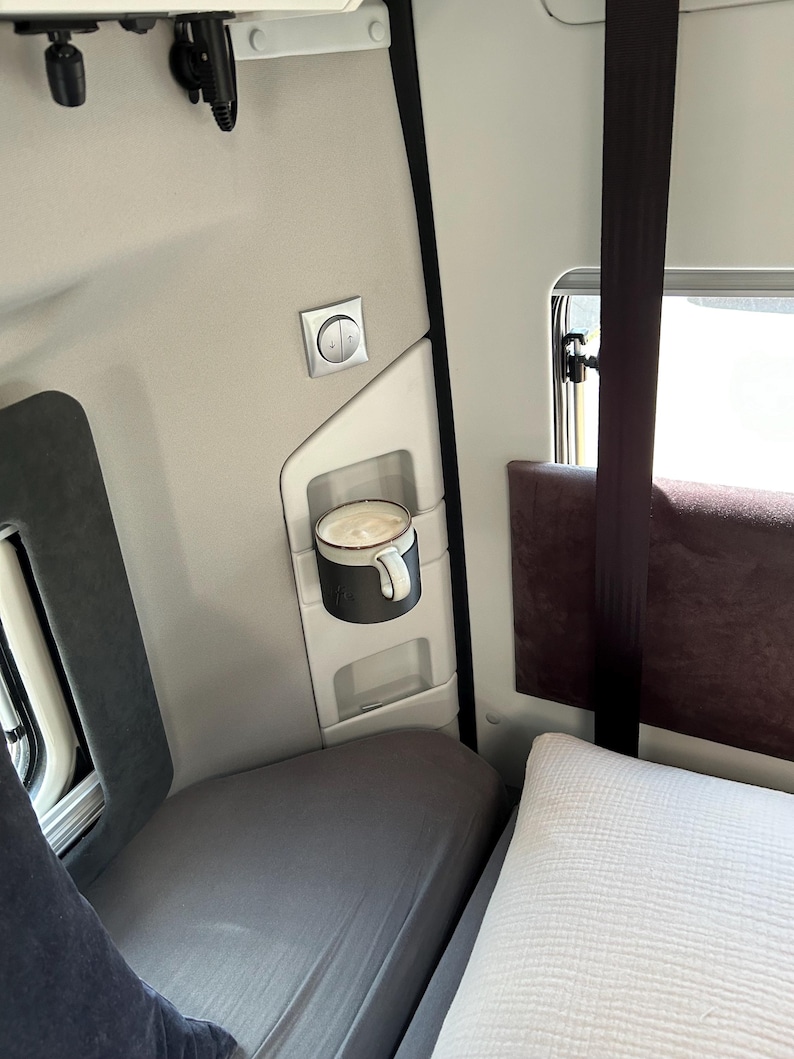 May include: A white ceramic mug with a black handle sits in a holder on the wall of a truck cab. The mug is filled with a light brown beverage. The cab has a gray and white interior with a window and a gray bed.