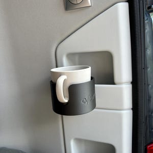 May include: A black cup holder with the word "van" embossed on it, holding a white ceramic mug. The cup holder is mounted on a gray surface.