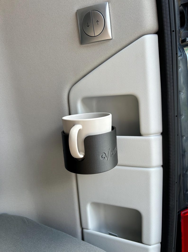 May include: A black cup holder with the word "van" embossed on it, holding a white ceramic mug. The cup holder is mounted on a gray surface.