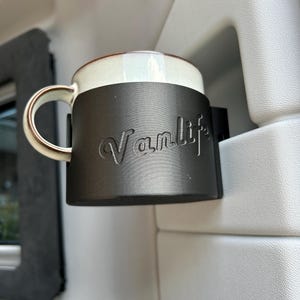 May include: A black cup holder with the word "Vanlife" embossed on it. A white ceramic mug is in the holder.