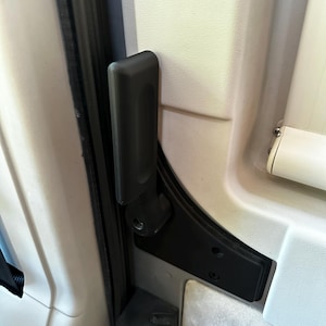 May include: A black plastic door handle with a silver metal hinge. The handle is attached to a white door panel.