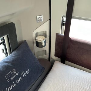 May include: A white bed with a blue pillow with the text "Twinni on Tour" embroidered on it. A white mug with a brown drink is in a holder on the wall. A white light switch is on the wall.