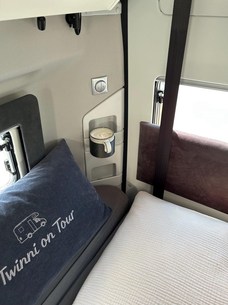 May include: A white bed with a blue pillow with the text "Twinni on Tour" embroidered on it. A white mug with a brown drink is in a holder on the wall. A white light switch is on the wall.