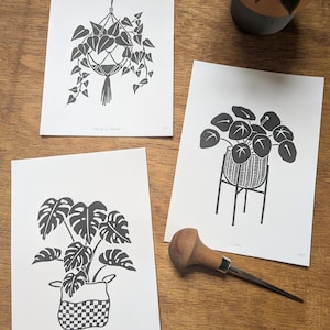 May include: Three black and white prints of houseplants in pots. The prints are on white paper with the plant names printed below each image. The prints are on a wooden surface.
