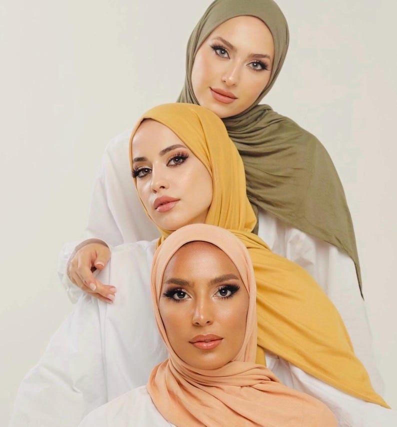 May include: Three women wearing headscarves in different colors: olive green, mustard yellow, and peach. The women are looking at the camera and smiling.
