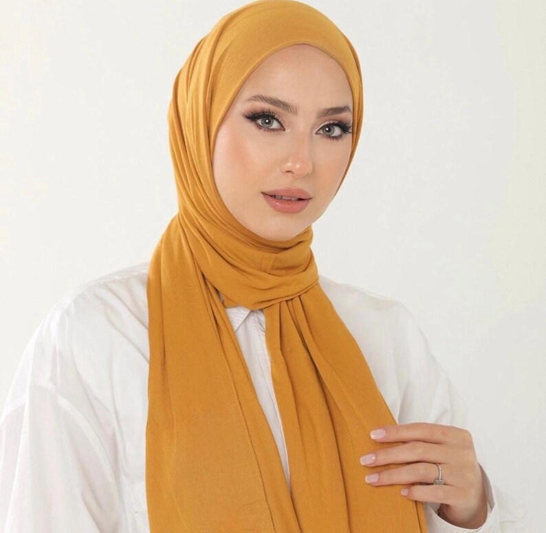 May include: A mustard yellow stretchy hijab made of soft fabric. The hijab is worn in a simple style with the ends draped down the front.