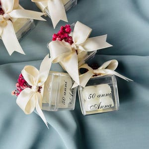 May include: Small clear boxes with white lids tied with ivory bows and decorated with small sprigs of pink flowers. Each box has a white label with black text that reads "50 anni Anna".