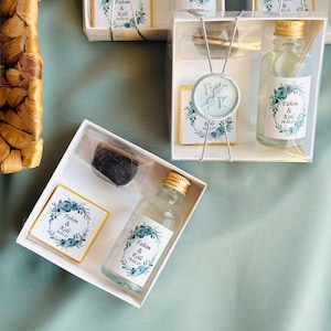 May include: A set of wedding favours in white boxes with blue and gold accents. Each box contains a small bottle of clear liquid with a label that reads "Fahim & Koli 16.02.21" and a small square box with a similar label.