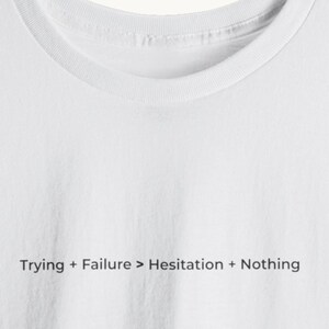 以下が含まれることがあります： 「Trying + Failure > Hesitation + Nothing」のテキストが黒でプリントされた白いクルーネックTシャツ。柔らかく快適な素材で作られています。