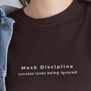 Könnte beinhalten: Ein dunkelbraunes T-Shirt mit dem weißen Text "Mock Discipline success loves being ignored". Das T-Shirt hat einen Rundhalsausschnitt und ist aus weichem Material. Auf der linken Seite des Bildes ist eine Jeansjacke zu sehen.