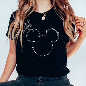 Puede incluir: Camiseta negra con un contorno blanco de la cabeza de Mickey Mouse, compuesto por estrellas y puntos. Un peque&ntilde;o hada est&aacute; dibujada encima de la cabeza, sosteniendo una varita. La camiseta es de estilo cuello redondo cl&aacute;sico.