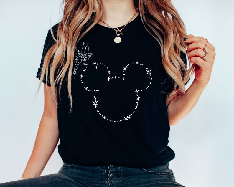 Puede incluir: Camiseta negra con un contorno blanco de la cabeza de Mickey Mouse, compuesto por estrellas y puntos. Un peque&ntilde;o hada est&aacute; dibujada encima de la cabeza, sosteniendo una varita. La camiseta es de estilo cuello redondo cl&aacute;sico.