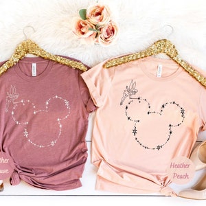Puede incluir: Dos camisetas, una color malva jaspeado y otra melocot&oacute;n jaspeado, cada una con un contorno de cabeza de Mickey Mouse hecho de estrellas y un hada. Las camisetas est&aacute;n colgadas en perchas doradas con lentejuelas.