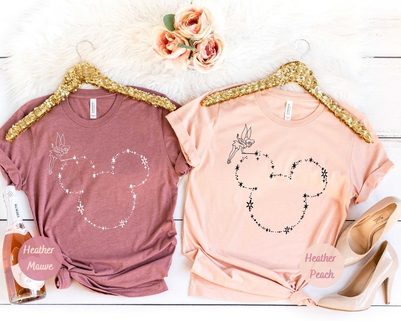 Puede incluir: Dos camisetas, una color malva jaspeado y otra melocot&oacute;n jaspeado, cada una con un contorno de cabeza de Mickey Mouse hecho de estrellas y un hada. Las camisetas est&aacute;n colgadas en perchas doradas con lentejuelas.