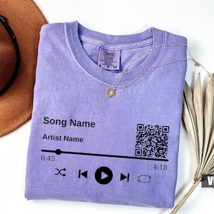 May include: A purple t-shirt with a music player design. The design includes a QR code, song name, artist name, play controls, and a timer. The text on the shirt reads "Song Name", "Artist Name", "0:45", and "4:18".