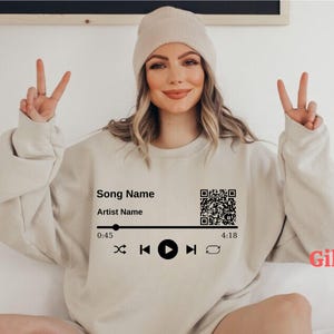 May include: Beige sweatshirt with a music player graphic. The graphic includes the text "Song Name" and "Artist Name", a QR code, and playback controls. The sweatshirt is a Gildan brand.