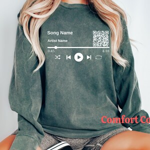 May include: A green sweatshirt with a white graphic of a music player with a song title, artist name, a QR code, and play controls. The text on the graphic reads "Song Name" and "Artist Name". The time on the graphic reads "0:45" and "4:18". The text "Comfort Colors" is printed on the bottom right corner of the sweatshirt.