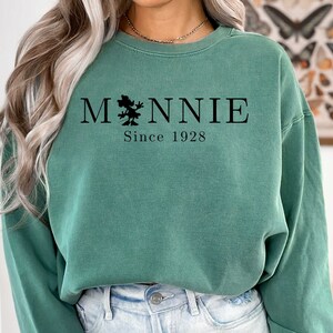 K&ouml;nnte beinhalten: Ein gr&uuml;ner Pullover mit einer schwarzen Grafik eines Comic-Charakters und dem Text "MNNIE Since 1928".