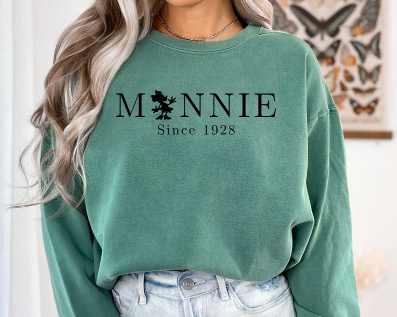 K&ouml;nnte beinhalten: Ein gr&uuml;ner Pullover mit einer schwarzen Grafik eines Comic-Charakters und dem Text "MNNIE Since 1928".