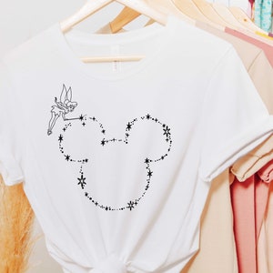 Puede incluir: Camiseta blanca con un dibujo lineal negro de Campanilla y una cabeza de Mickey Mouse hecha de estrellas y puntos. La camiseta est&aacute; colgada en una percha de madera con otras camisetas en el fondo.