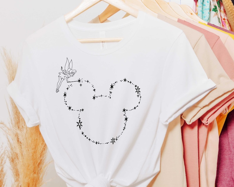 Puede incluir: Camiseta blanca con un dibujo lineal negro de Campanilla y una cabeza de Mickey Mouse hecha de estrellas y puntos. La camiseta est&aacute; colgada en una percha de madera con otras camisetas en el fondo.